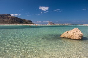 Ett turkosblått hav med en stor klippa i förgrunden och en strand med människor i bakgrunden.