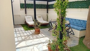Eine Terrassenfläche mit einer Pergola, zwei Liegestühlen, einer blauen gepolsterten Bank und Kübelpflanzen.