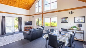 Ein geräumiges Wohnzimmer mit hohen Decken, einem Fernseher, einer Couch, einem Esstisch mit Stühlen und großen Fenstern mit Blick auf Grünflächen.