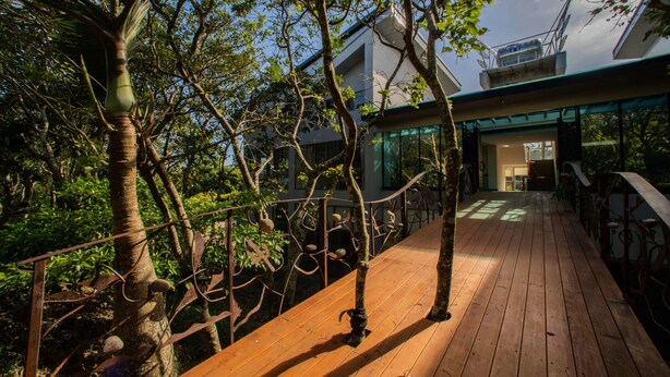 A wooden deck with a metal railing, surrounded by trees and leading to a modern building with large windows. - TopTai B&B (Hengchun)