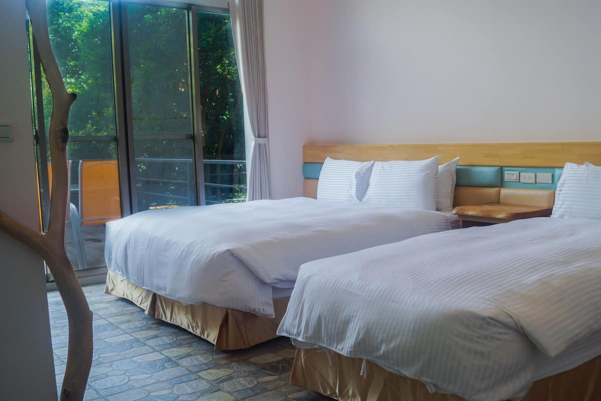 A hotel room with two beds, a wooden headboard, a nightstand, and a view of greenery through the window.