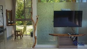 A modern living room with a large flat-screen TV mounted on a wooden console, a glass partition, and a view of greenery outside. - TopTai B&B (Hengchun)