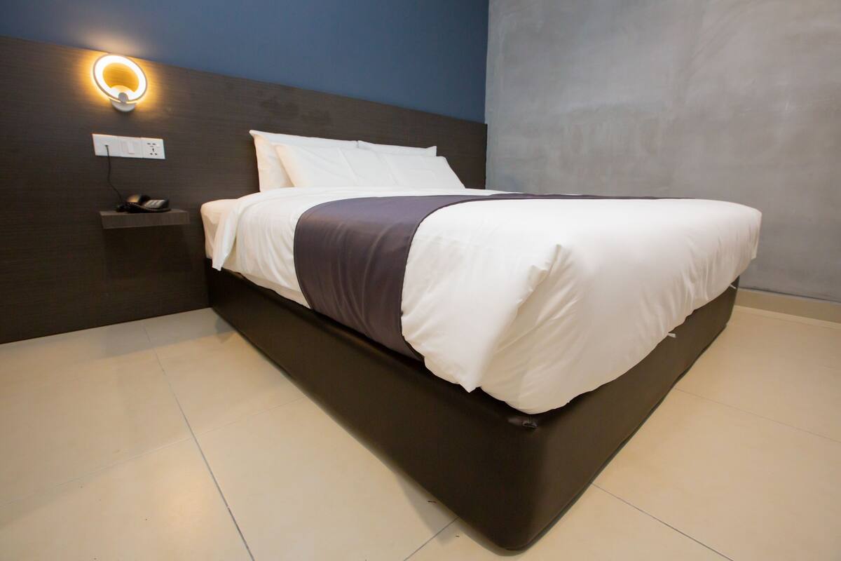 A modern hotel room with a large bed, a bedside table with a phone, and a wall-mounted light fixture.