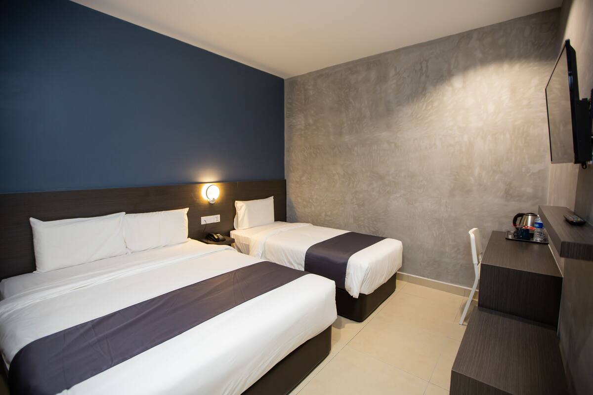 A hotel room with two beds, a desk, a chair, a TV, and a wall-mounted light.
