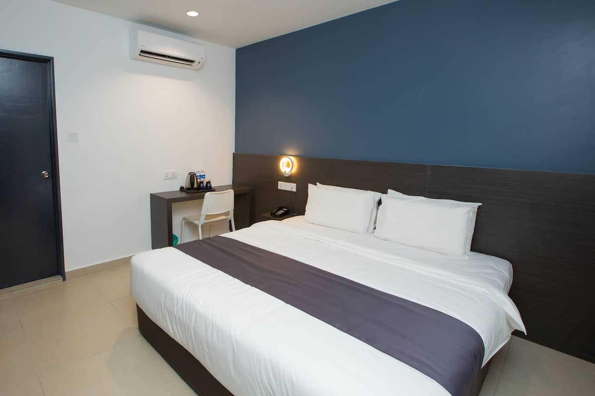 A hotel room with a large bed, a desk with a chair, a nightstand, and a wall-mounted lamp.