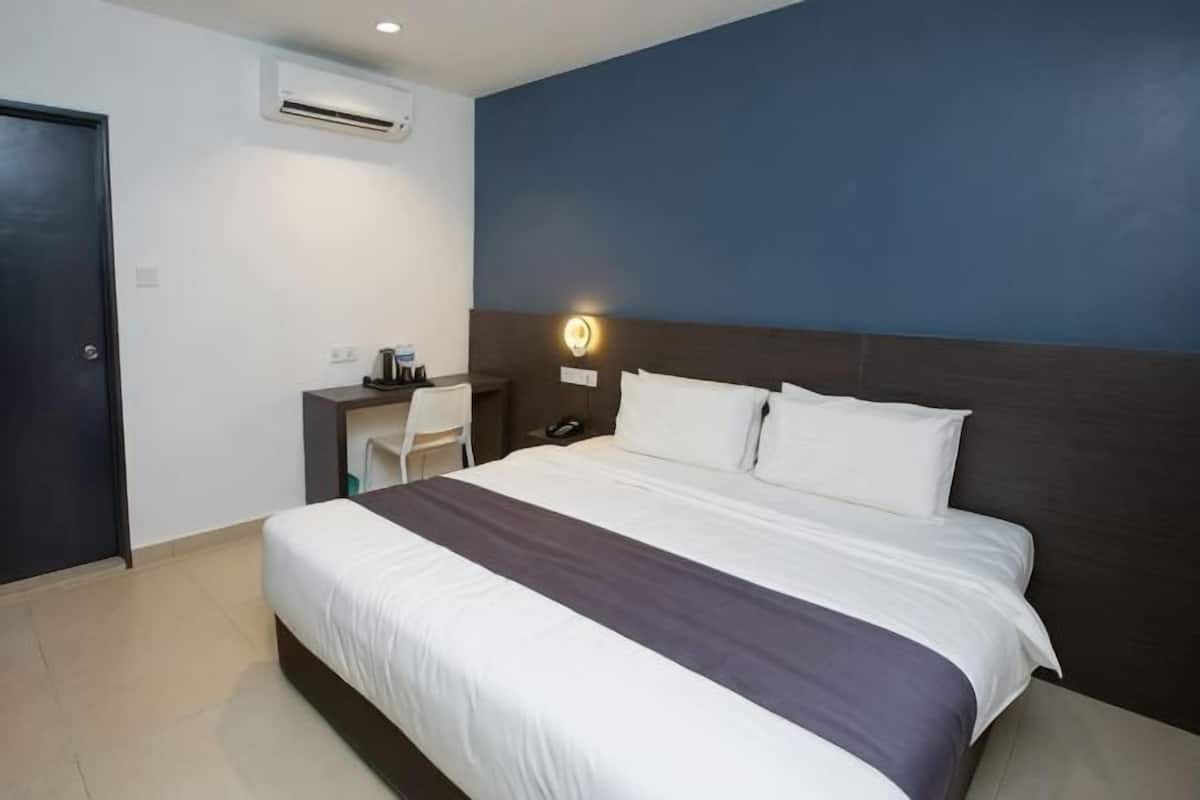 A hotel room with a bed, bedside table, chair, desk, and a wall-mounted air conditioner.