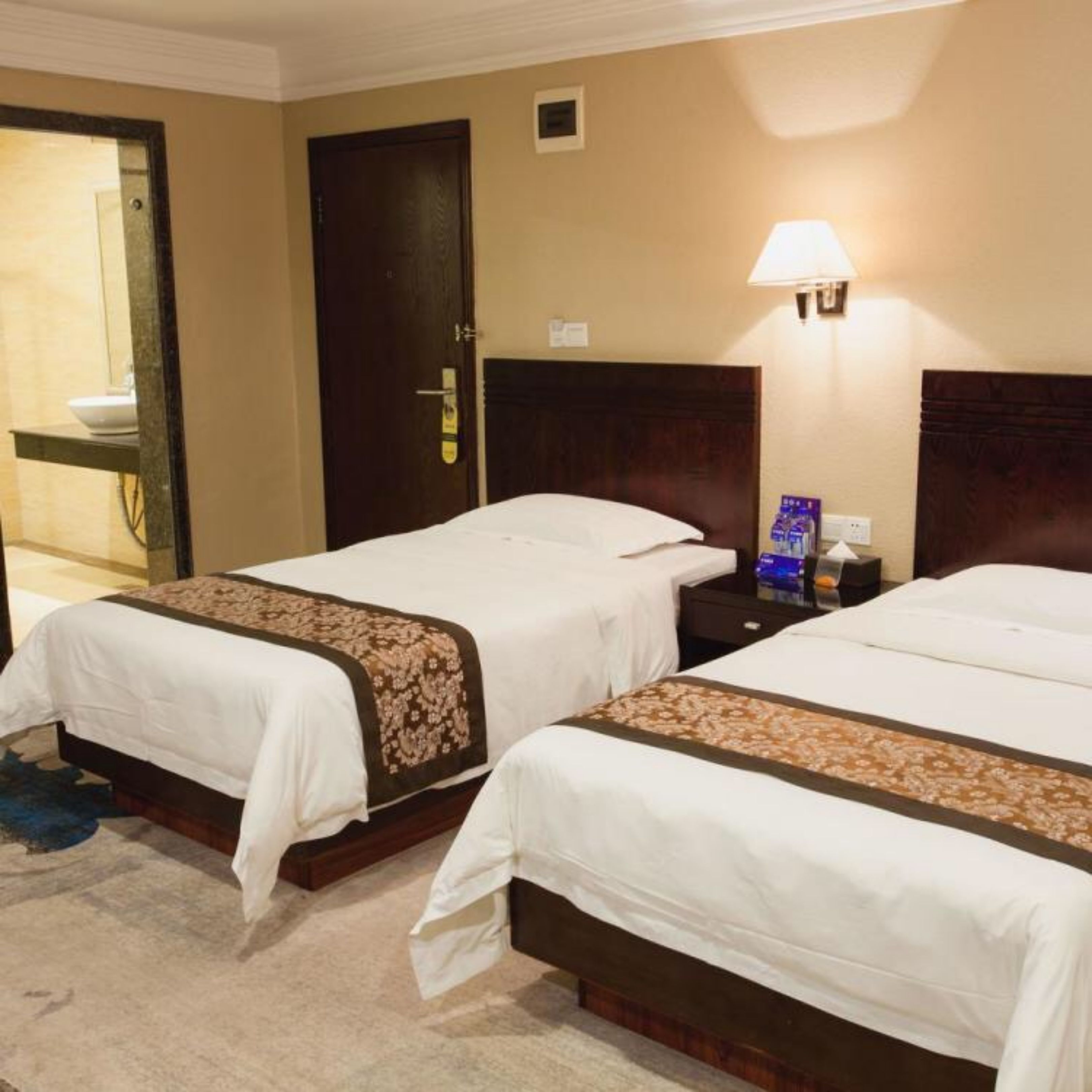 A hotel room with two beds, a nightstand with a lamp, a door, and a small window.