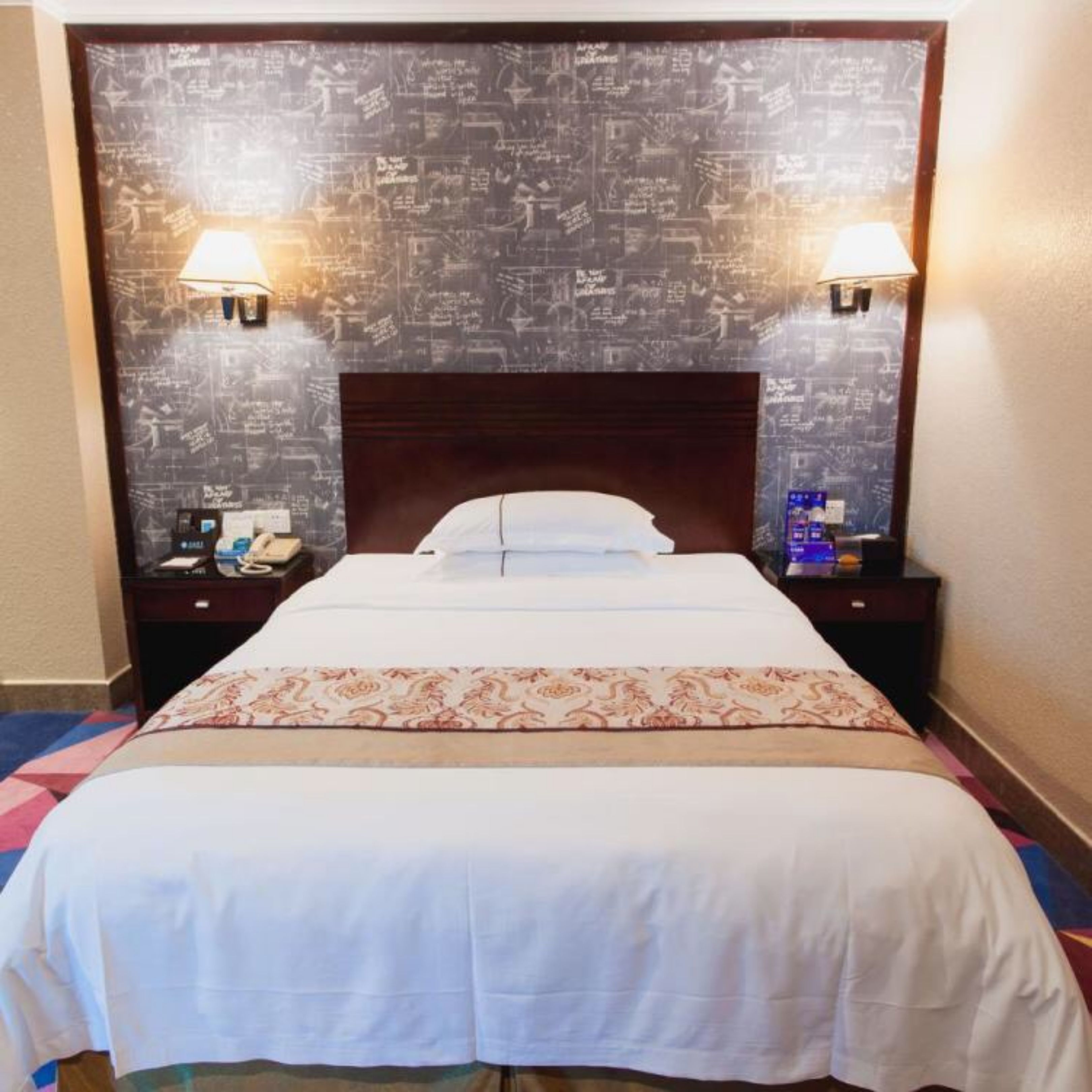 A hotel room with a large bed, two bedside tables, a headboard with a chalkboard design, and two wall-mounted lamps.