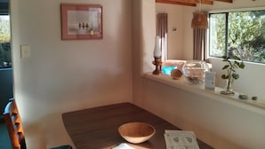 A cozy room with a wooden dining table, a framed picture on the wall, a window with a view of trees, and a shelf with decorative items.