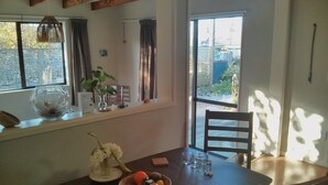 A dining area with a wooden table, chairs, a bowl of fruit, a vase with flowers, and a window with curtains.