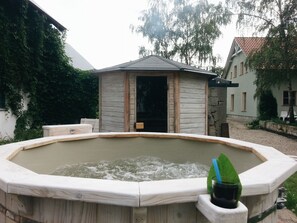 A wooden hot tub with a wooden deck surrounding it, set in a garden with a building in the background.