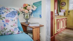 Een kamer met een bed, een bureau met een bloevas, een schilderij en een keukenhoek met een magnetron.