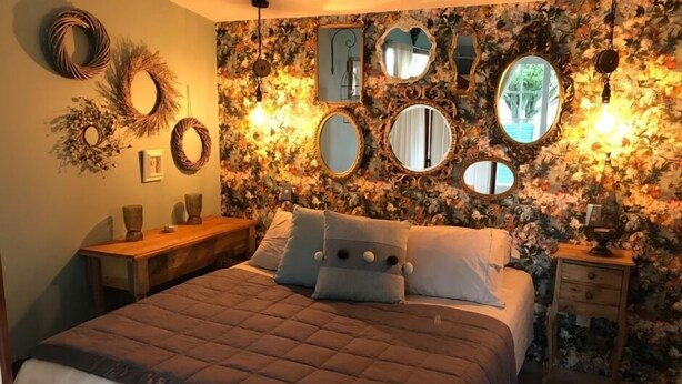 A bedroom with a floral wallpaper, a bed with a brown quilt, multiple mirrors, and a nightstand with a lamp. - Kokako Lodge Wanganui (Wanganui Township)