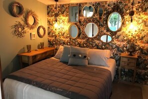 Un dormitorio con papel tapiz floral, una cama con colcha marrón, varios espejos y una mesita de noche con lámpara.