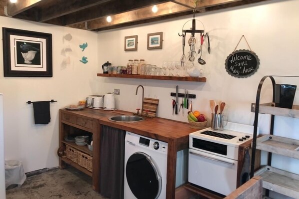 Cocina compacta con lavadora, microondas y estufa. Hay estantes con frascos y vasos, y un cartel de bienvenida.