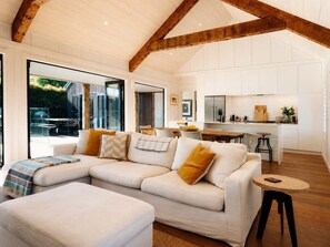A modern living room with a sectional sofa, a small round table, and a kitchen area with white cabinets and wooden beams.