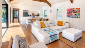 A spacious living area with a white sofa, ottoman, and a dining table with chairs. A kitchen is visible in the background. - Blackpool Boathouse - Pool & Spa & Walk to Oneroa (Oneroa)