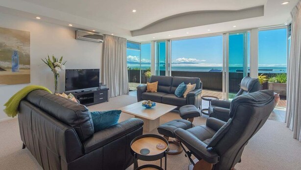 Ein modernes Wohnzimmer mit großem Fernseher, bequemen Sitzmöglichkeiten und einem Balkon mit Meerblick.