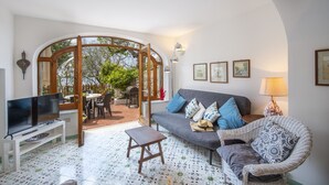 Een woonkamer met een televisie, bank en rieten stoel, een eethoek met tafel en stoelen, en uitzicht op groen via een glazen deur.