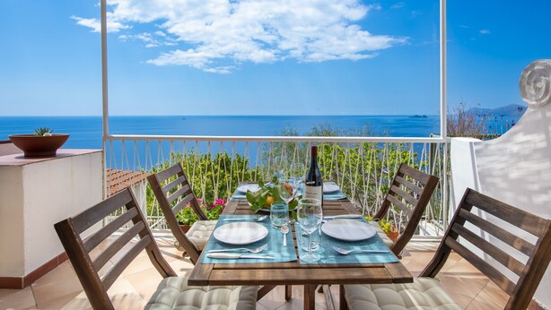 Een eettafel gedekt voor twee personen met uitzicht op zee, een fles wijn en een potplant op een balkon.