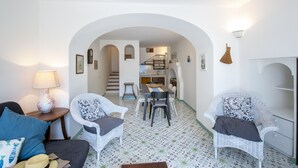 Een gezellige zithoek met een bank, twee witte rieten stoelen, een eettafel, en een keuken op de achtergrond.
