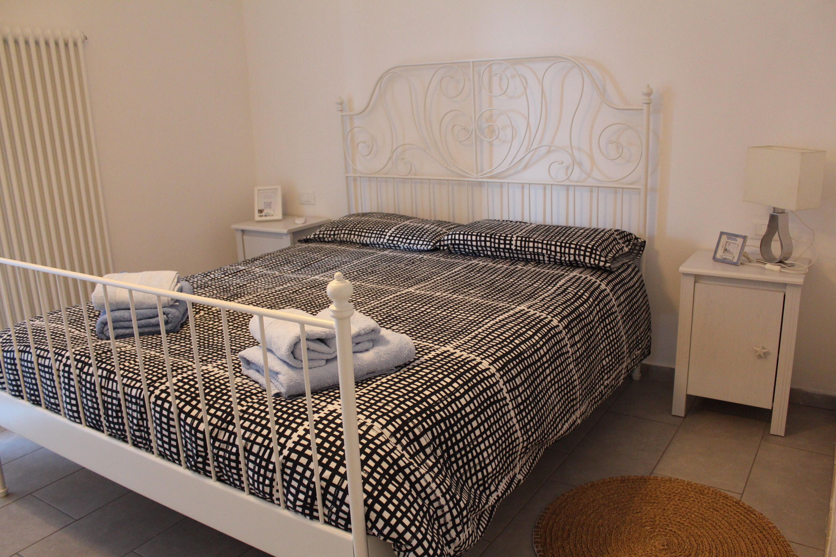 Een wit metalen bed met een bedspreide met een patroon en kussens, een nachtkastje met een lamp, en een klein vloerkleed.