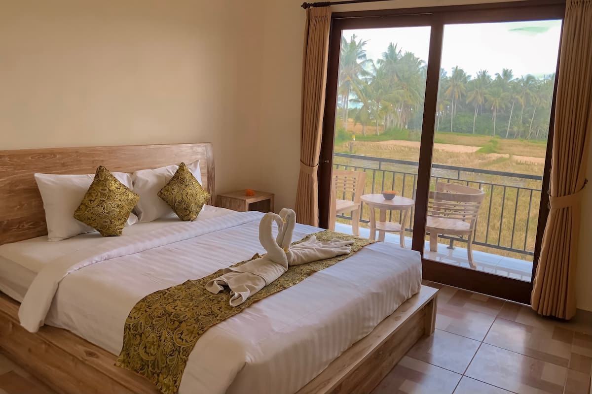 Ein Hotelzimmer mit einem großen Bett, einem hölzernen Kopfteil, zwei Zierkissen, einem Nachttisch mit einer Schale und Blick auf eine Palmenlandschaft durch eine Schiebetür.