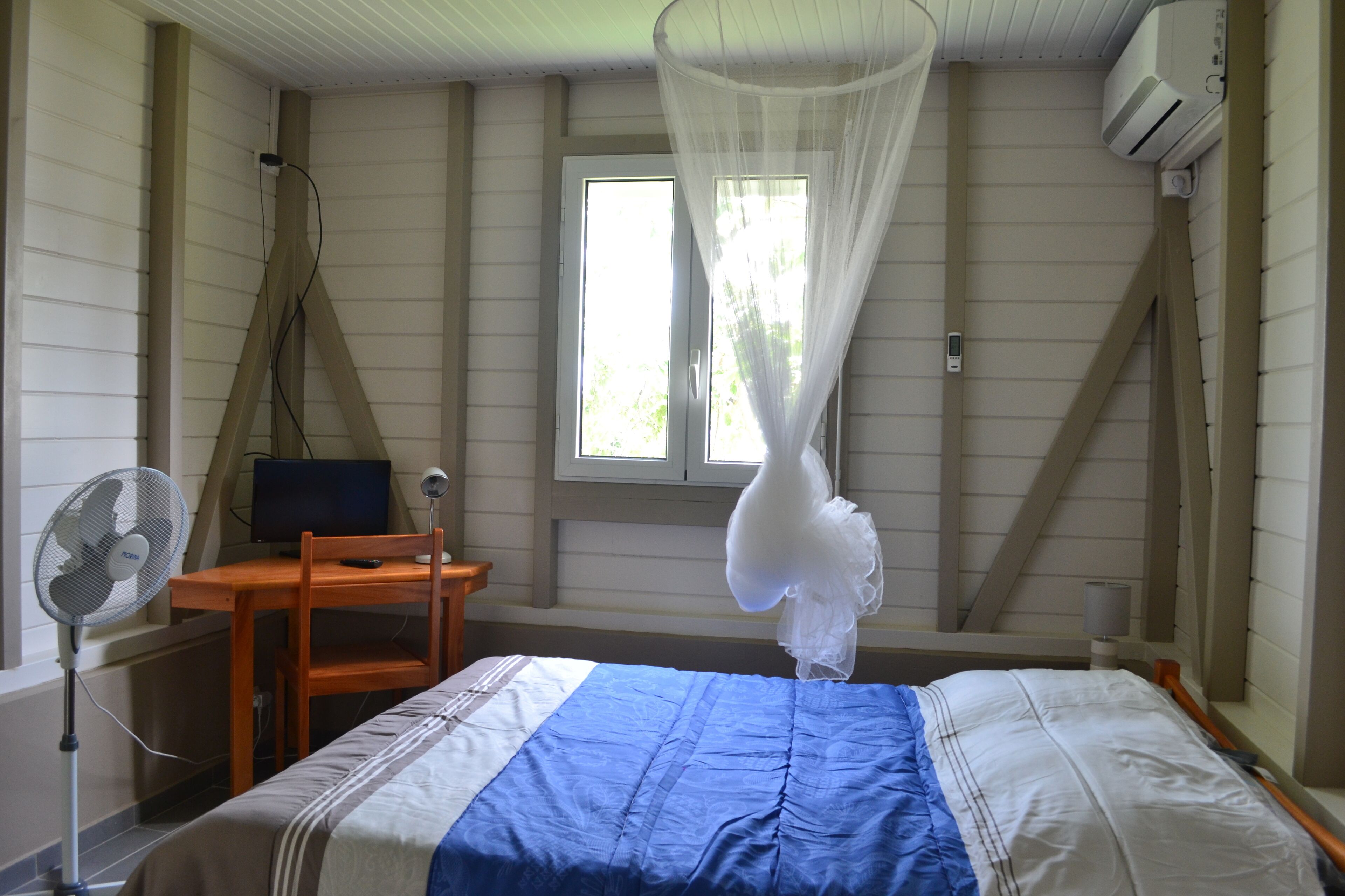Een slaapkamer met een bed, een bureau, een stoel, een ventilator en een raam met een witte gordijn.