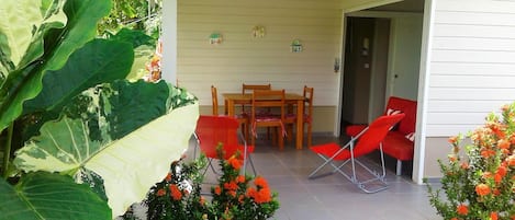 A covered patio with a wooden table and chairs, surrounded by large green leaves and orange flowers.