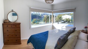 A bedroom with a large window, a wooden dresser, a bed with a blue blanket, and a mirror. - Cissy Bay holiday home (Elaine Bay)