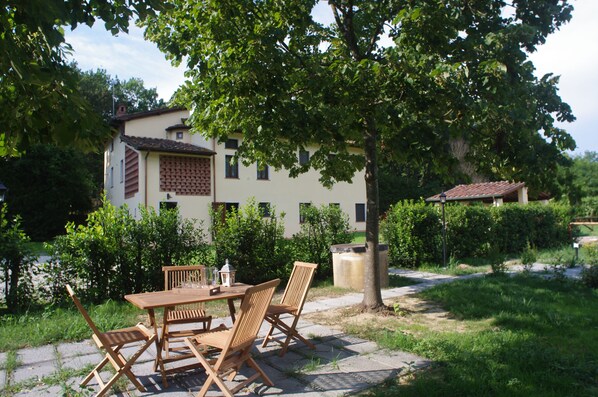 A patio with a wooden table and chairs, surrounded by greenery and a building with a tiled roof.