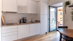 Et moderne køkken med hvide skabe, en bordplade, kaffemaskine og vask. Der er et træbord med stole, og en skydedør, der fører ud til en balkon.