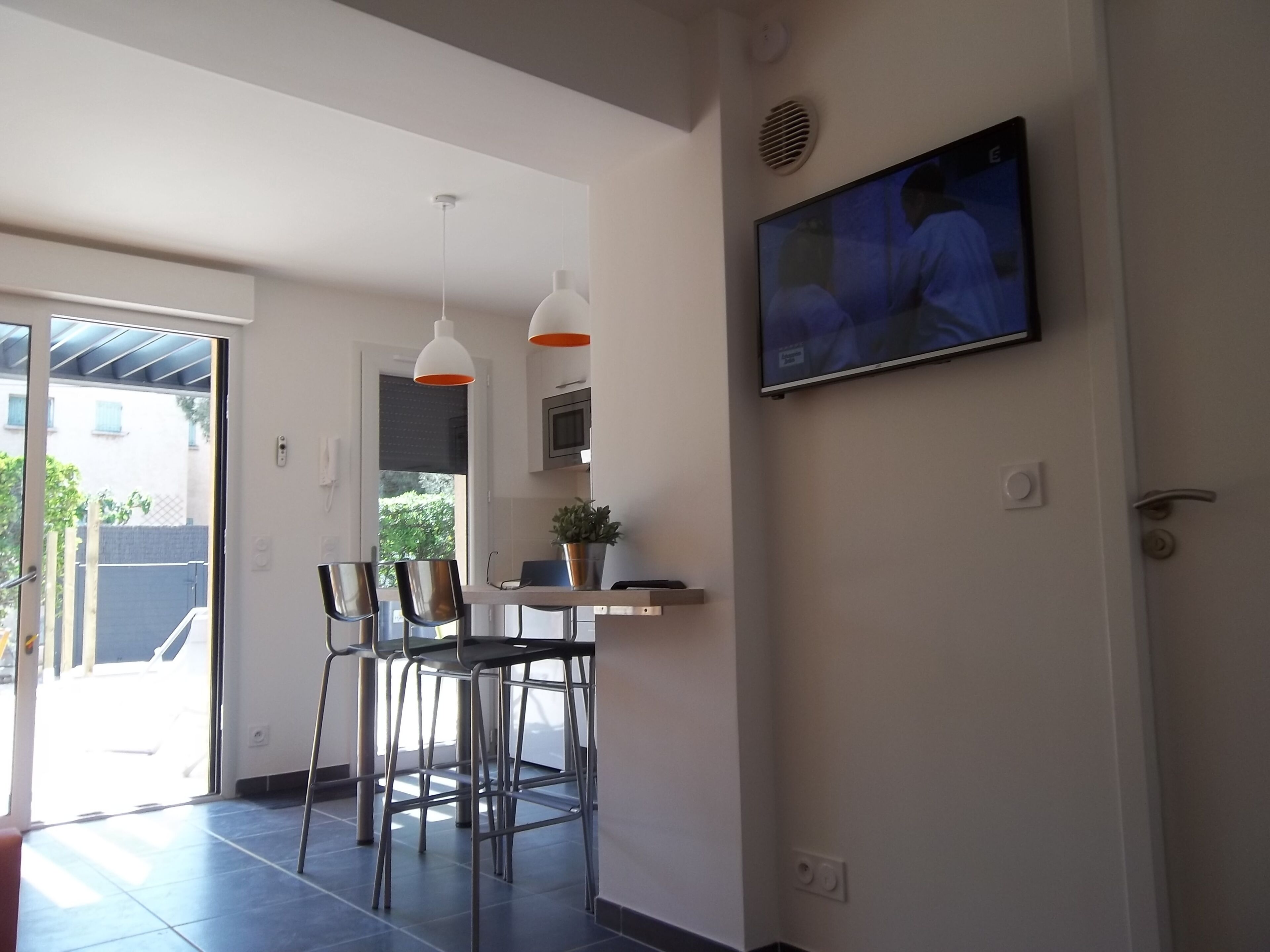 A modern kitchen with a dining area, a TV mounted on the wall, and a sliding glass door leading to a patio.