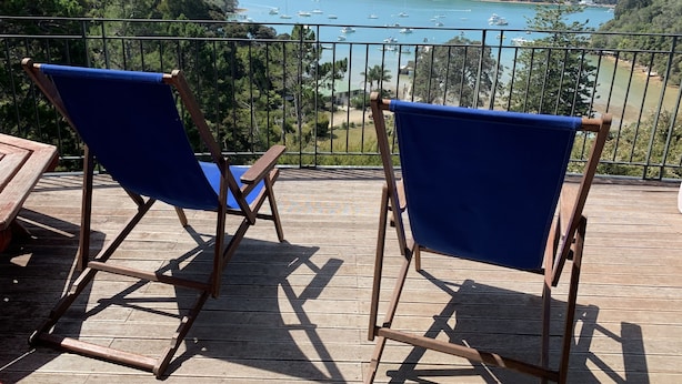 Two blue folding chairs on a wooden deck overlooking a bay with boats and hills. - Peaceful Ocean facing Island Escape (Kawau Island)