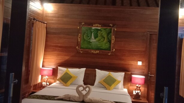 A wooden-framed bed with white bedding, two pillows with green and yellow accents, and a heart-shaped towel arrangement. A framed painting of a green landscape hangs above the bed. - Svaha Nusa Guest House (Lembongan Island)