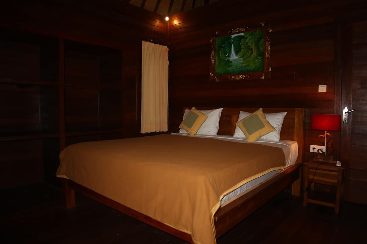 A large bed with a wooden headboard, a nightstand with a red lamp, and a painting of a waterfall on the wall.