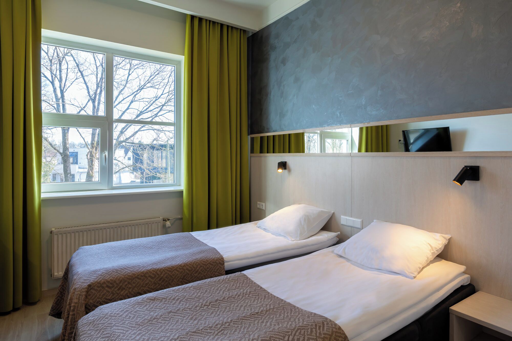 A hotel room with two beds, a window with curtains, a radiator, and a wall-mounted TV.