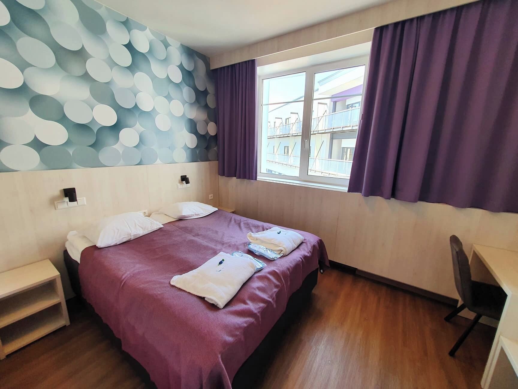 Une chambre d’hôtel comprenant un lit, un bureau, une chaise, une fenêtre avec des rideaux et un mur orné d’un motif circulaire.