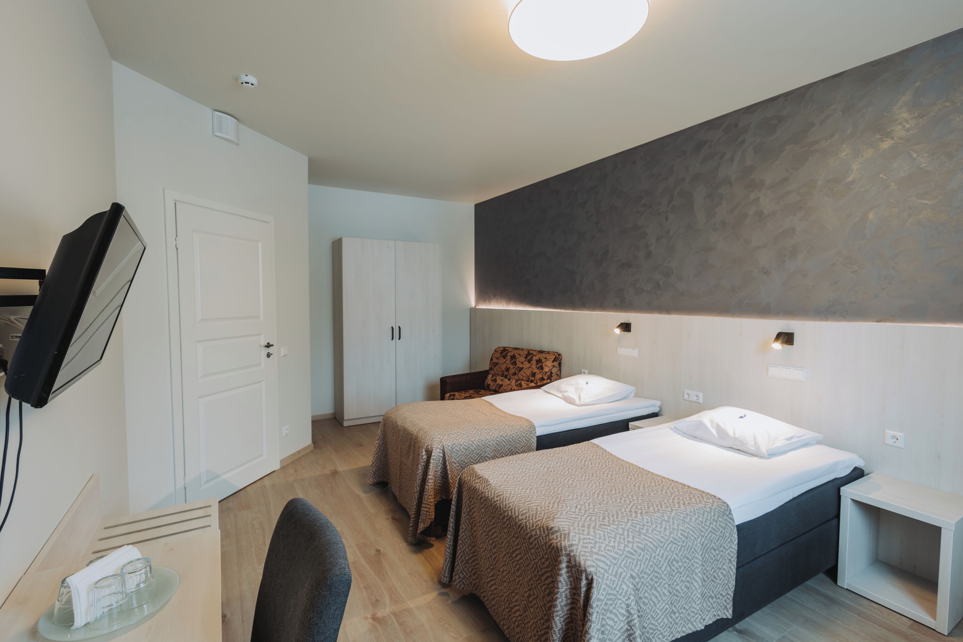 A modern hotel room with two single beds, a desk, a chair, a wardrobe, and a television.