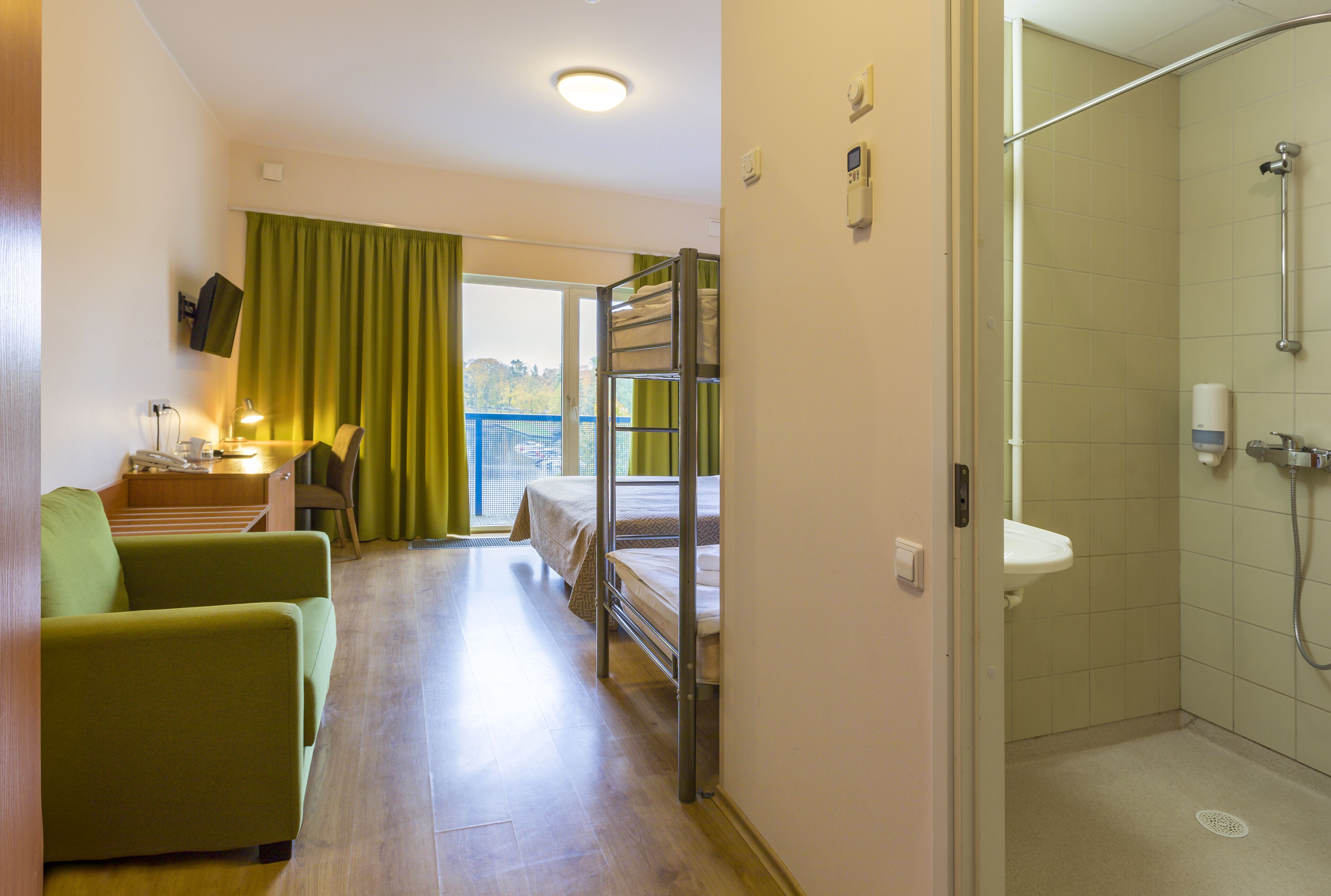 A hotel room with a bunk bed, a desk with a lamp, a green armchair, and a shower area.