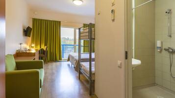 A hotel room with a bunk bed, a desk with a lamp, a green armchair, and a shower area.