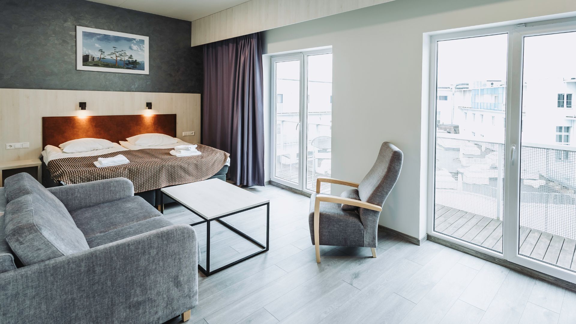 A modern hotel room with a bed, a sofa, a chair, a small table, and large windows offering a view of the city.