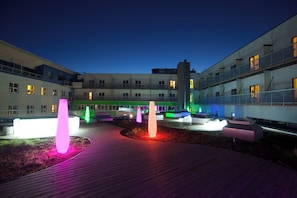 Un patio con arquitectura moderna, una terraza de madera y esculturas iluminadas.