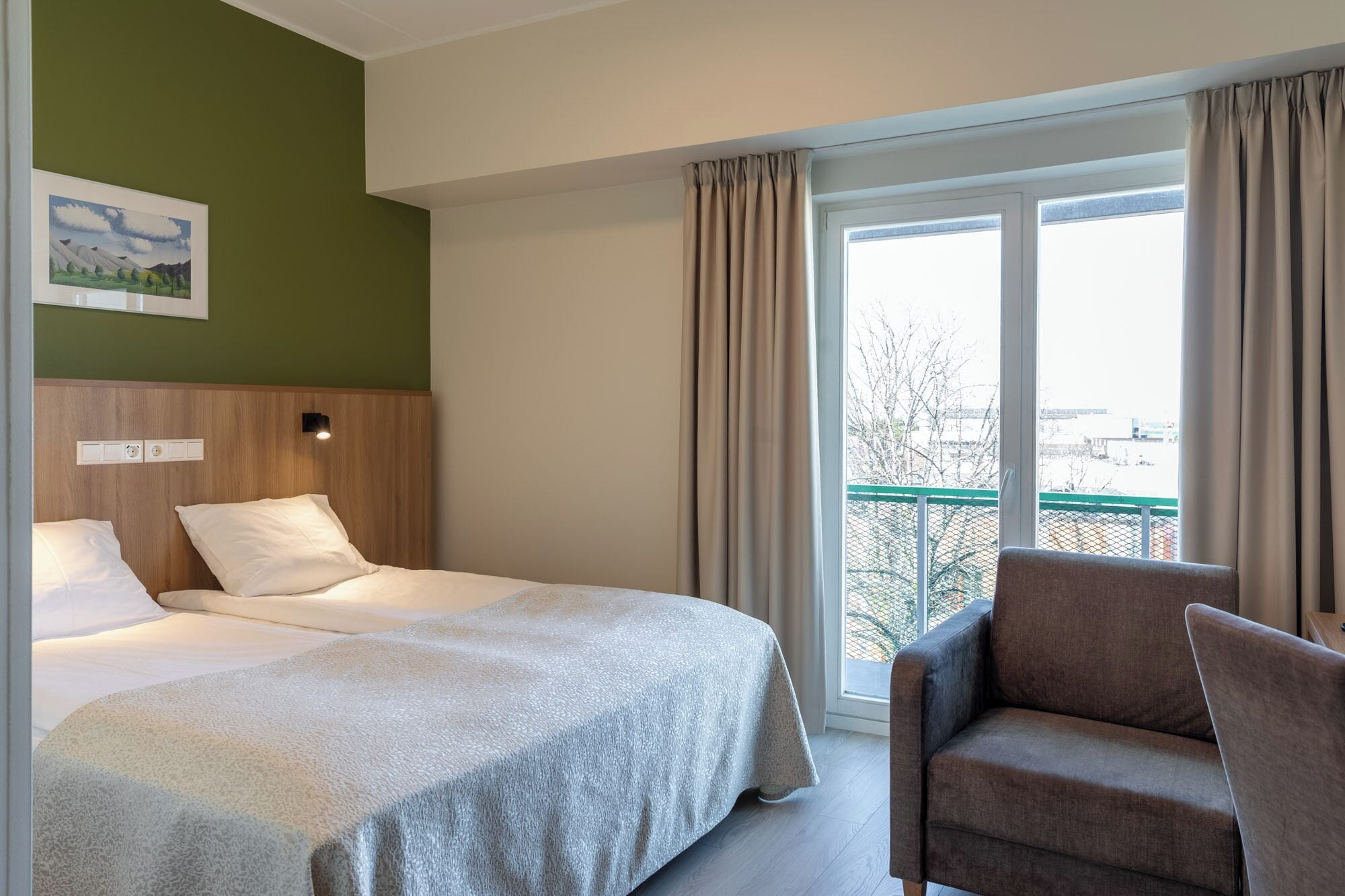 A hotel room with a bed, a chair, a window with curtains, and a wall-mounted light.