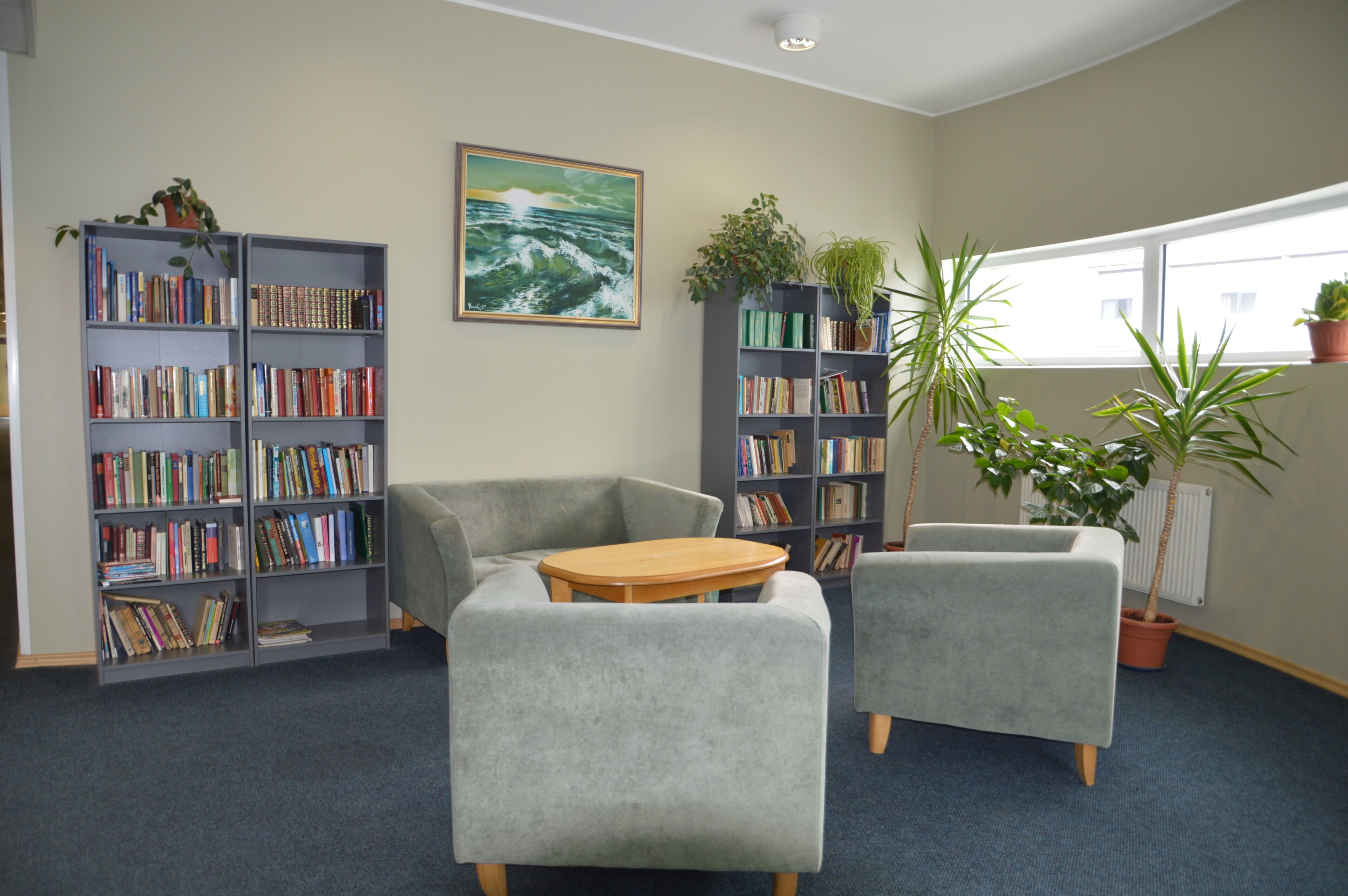 A library with two grey armchairs, a wooden table, and bookshelves filled with books.