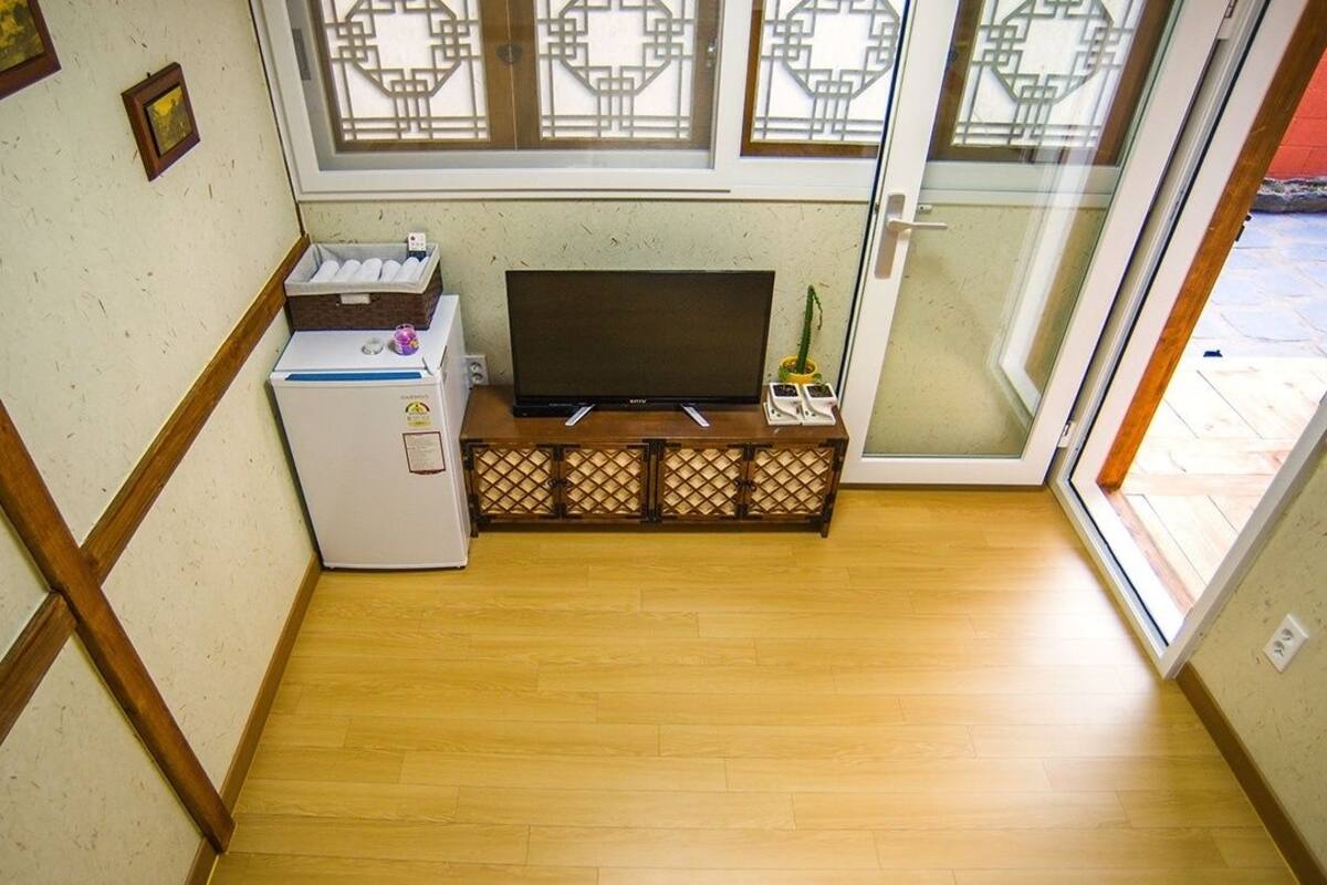 Habitación compacta con piso de madera, televisor sobre un bajo mueble, refrigerador pequeño y puerta corrediza de vidrio que da a un espacio al aire libre.