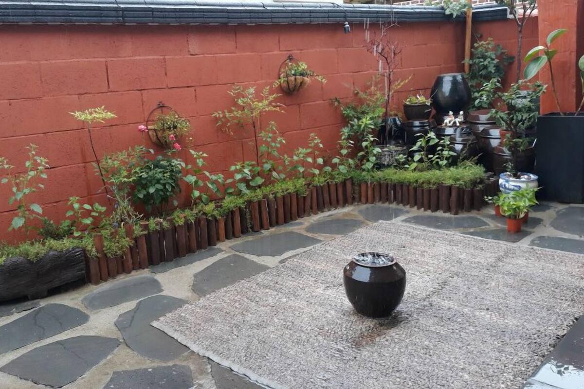 Un patio con suelo de piedra, una maceta negra decorativa en el centro, plantas en macetas y una pared roja.