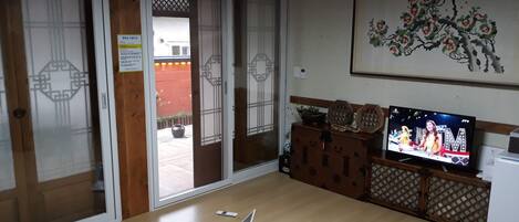 Una habitación con piso de madera, un televisor sobre un soporte, una computadora portátil en el suelo y una puerta con un panel decorativo.