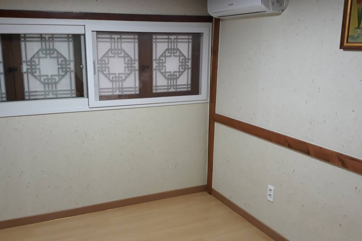 Una habitación con piso de madera, pared blanca con un panel decorativo y ventana con diseño geométrico.
