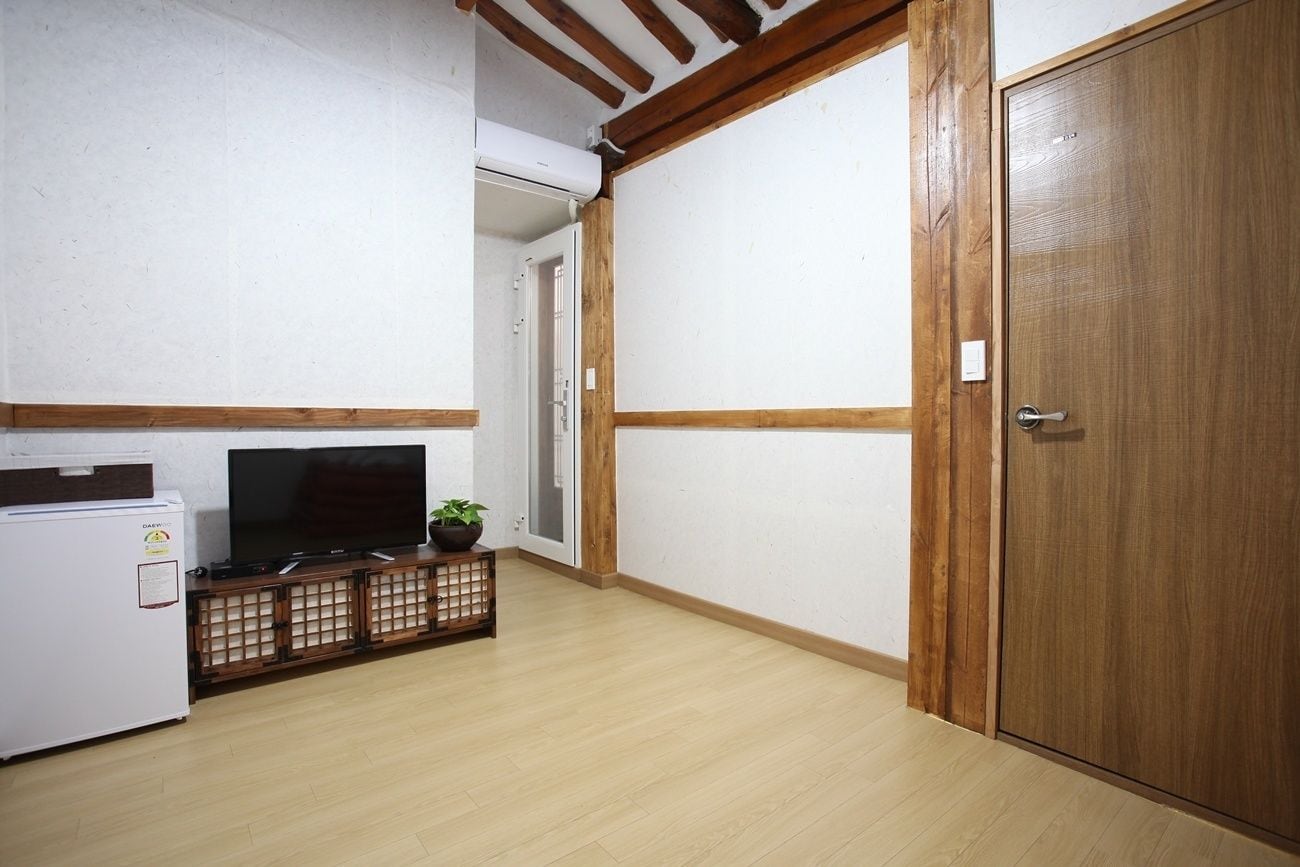 Quarto com piso de madeira, uma geladeira branca, TV de tela plana em um suporte de madeira e porta de madeira.
